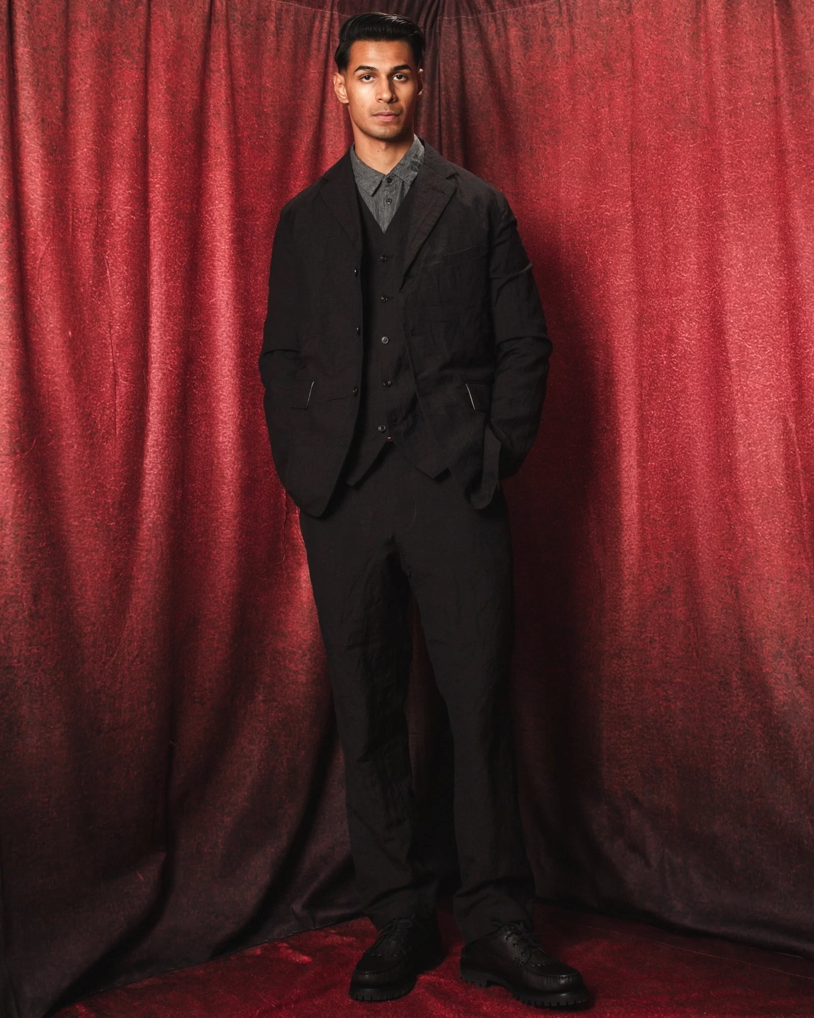 Sagar Shabbir — black tailored jacket and waistcoat over grey shirt, red curtain studio